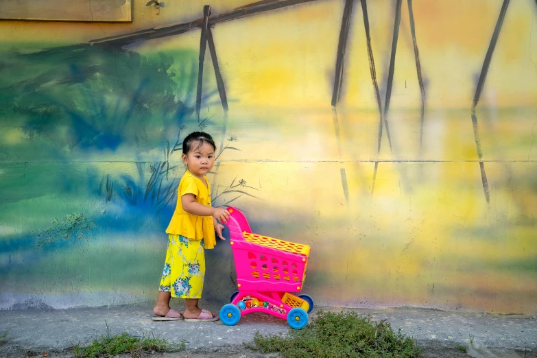 a little girl standing in front of a wall with a pink shopping cart, a picture, inspired by Pacita Abad, pexels contest winner, yellow colours, bali, avatar image, 2 years old