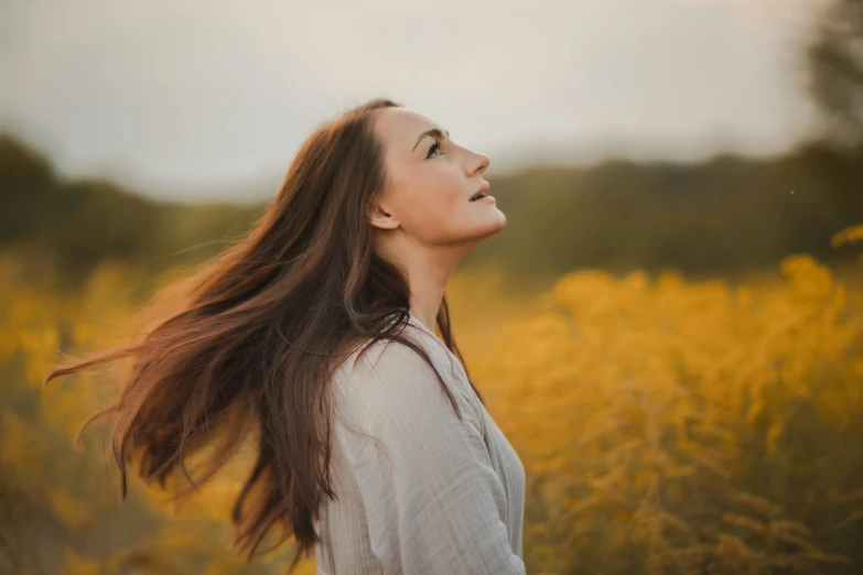 a woman standing in a field of yellow flowers, by Emma Andijewska, pexels contest winner, long flowing brown hair, relaxed. gold background, looking from side, face looking skyward