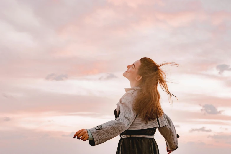 a woman standing on top of a lush green field, pexels contest winner, happening, orange / pink sky, windy floating hair!!, she is smiling and excited, looking across the shoulder