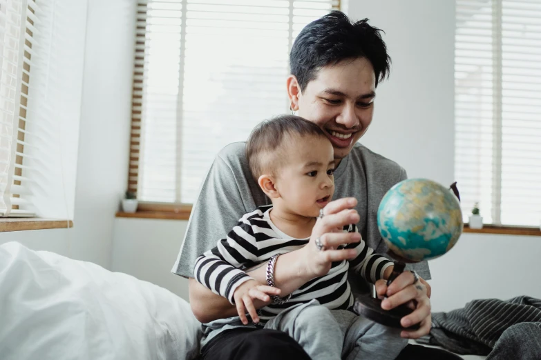 a man sitting on top of a bed holding a baby, pexels contest winner, inside a globe, asian male, with a kid, thumbnail