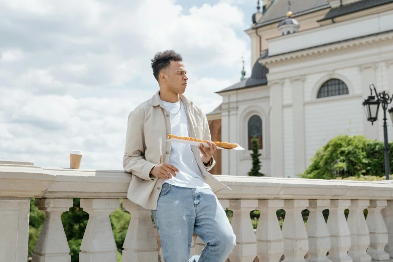 a man sitting on a ledge in front of a building, holding a baguette, millennial vibes, standing near a castle, kārlis padegs