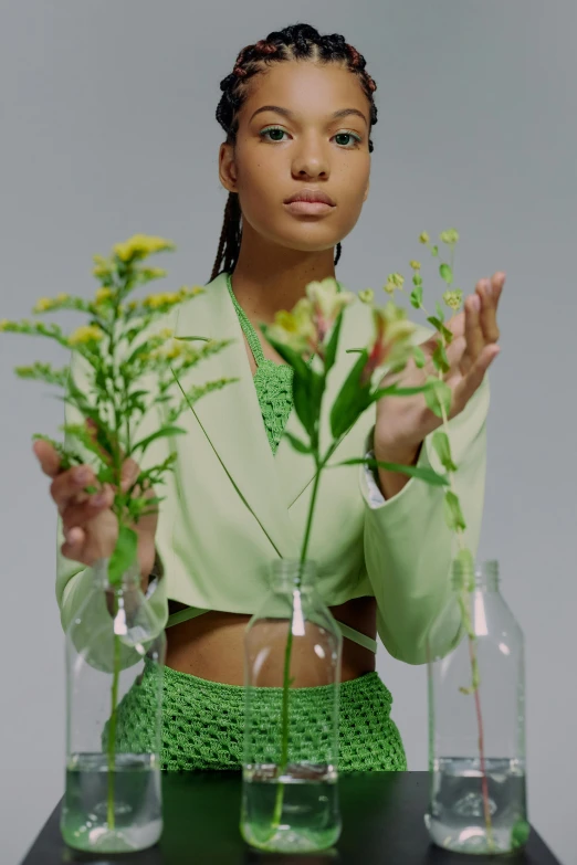 a woman standing in front of three vases filled with flowers, an album cover, trending on pexels, model is wearing techtical vest, plants in scientific glassware, wearing green clothing, willow smith young