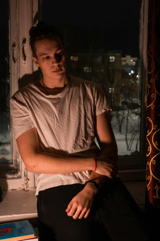 a man sitting on a window sill in front of a window, an album cover, inspired by Nan Goldin, pexels contest winner, alexey gurylev, he looks like tye sheridan, ( ( theatrical ) ), low lights