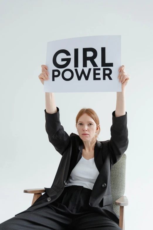 a woman sitting in a chair holding up a sign, trending on pexels, feminist art, power girl, on a gray background, hr ginger, superpowers