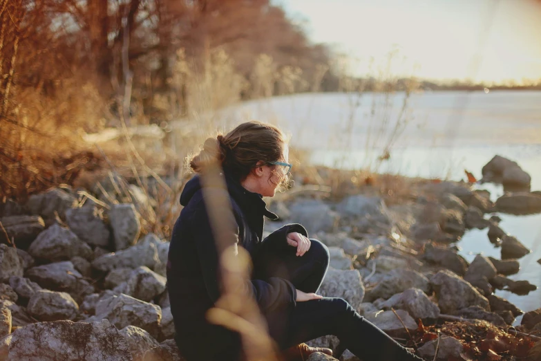 a woman sitting on a rock next to a body of water, by Emma Andijewska, girl with glasses, winter sun, unfocused, fall season