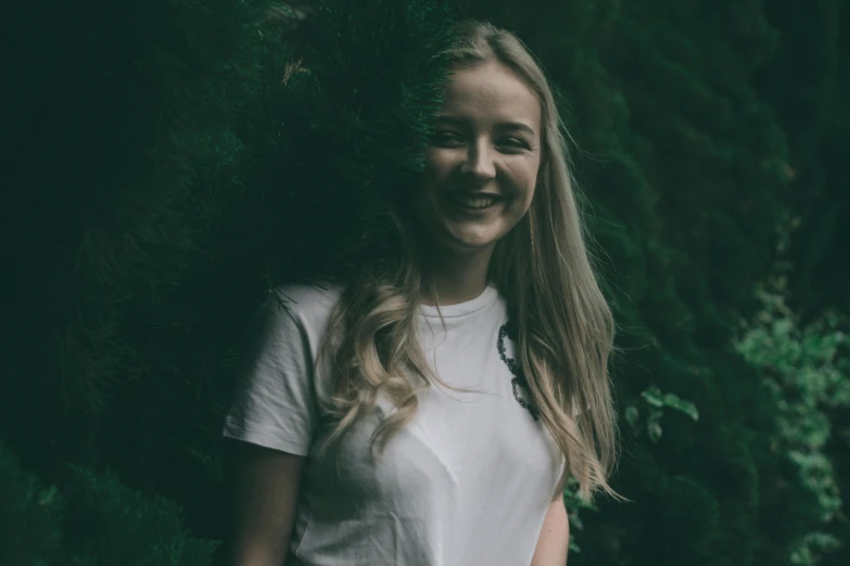 a young woman standing in front of a tree, pexels contest winner, slightly awkward smile, a girl with blonde hair, dressed in a white t shirt, avatar image