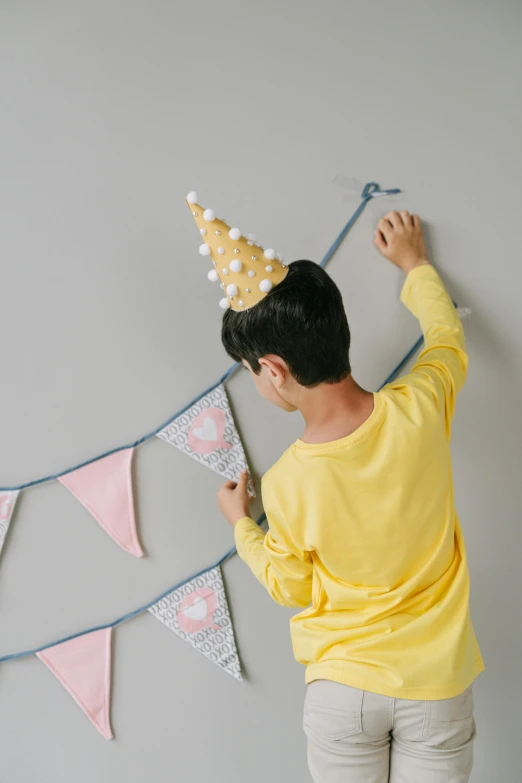 a person hanging a party hat on a wall, wearing a light grey crown, colors: yellow, kids, bedhead