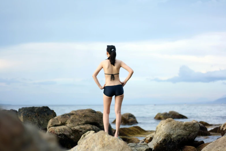 a woman standing on top of a rock next to the ocean, inspired by Liu Haisu, minimalism, sport bra and dark blue shorts, centered full body rear-shot, profile image, trending photo