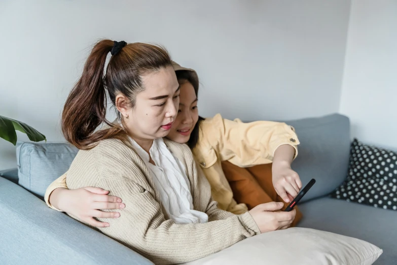 two women sitting on a couch looking at a cell phone, a cartoon, trending on pexels, hugging, asian human, background image, colour photograph