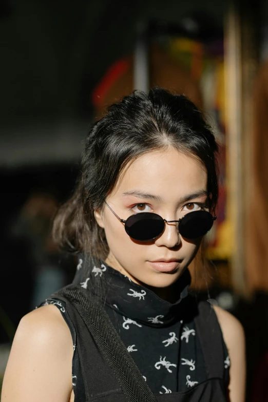a woman standing with a cell phone in her hand, trending on unsplash, photorealism, spiked collar sunglasses, ethnicity : japanese, black turtle neck shirt, south east asian with round face