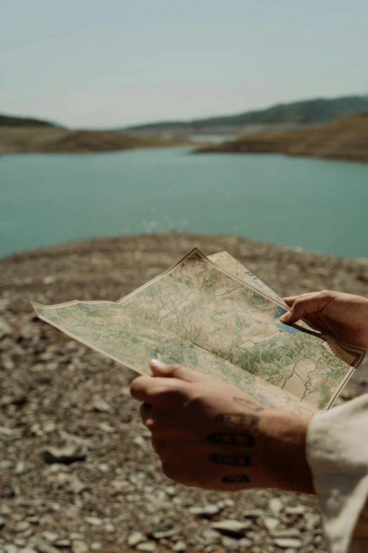 a person holding a map near a body of water, rugged, we go, brown, features