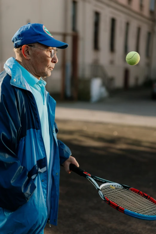 a man holding a tennis racquet next to a tennis ball, a portrait, by Jan Tengnagel, unsplash, an 80 year old man, wearing blue jacket, wearing sunglasses and a cap, finland