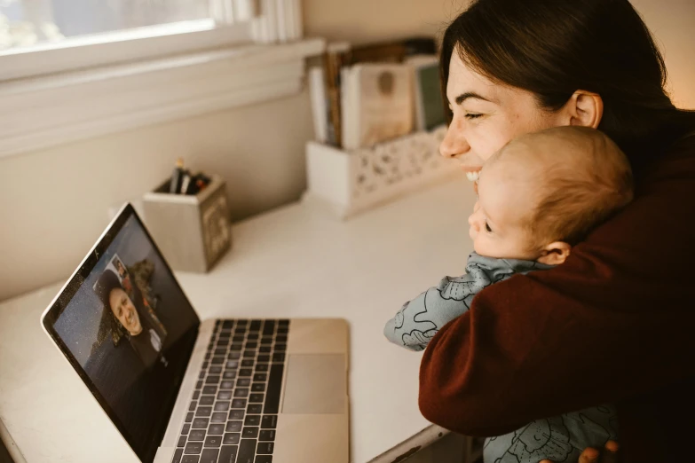 a woman holding a baby in front of a laptop, by Julia Pishtar, pexels contest winner, animated, a handsome, brown, webcam