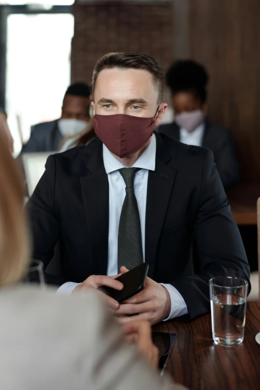 a man in a suit sitting at a table with a mask on his face, bringing people together, maroon, model is wearing techtical vest, stockphoto