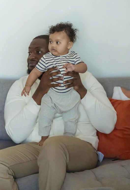 a man sitting on a couch holding a baby, inspired by Paul Georges, pexels contest winner, kevin hart, gilleard james, tall and strong, loosely cropped