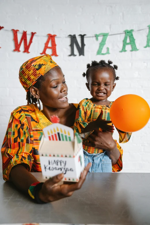 a woman holding a child while sitting at a table, an album cover, by Ingrida Kadaka, pexels contest winner, black arts movement, colorful paper lanterns, birthday wrapped presents, at a birthday party, african