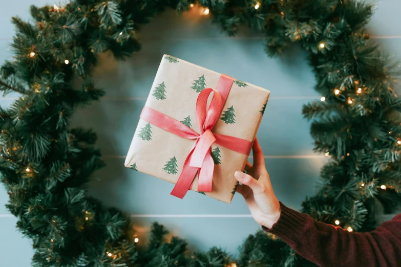 a person holding a wrapped gift in front of a christmas wreath, by Alice Mason, pexels contest winner, packaging, retro style ”, background image, small