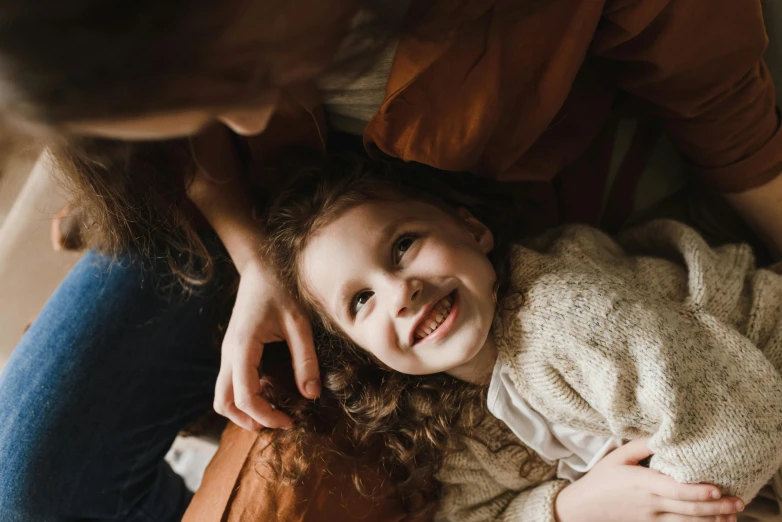 a woman sitting on top of a couch next to a little girl, by Emma Andijewska, pexels contest winner, brown curly hair, smiling down from above, warm coloured, thumbnail