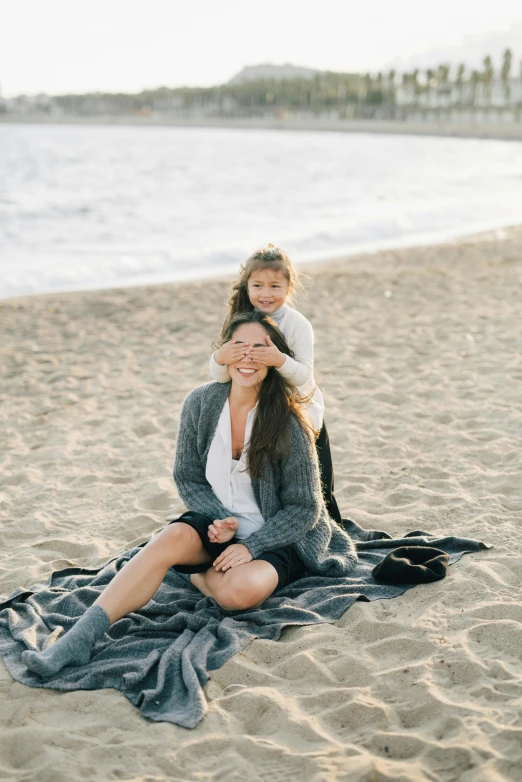 a woman sitting on top of a blanket on top of a beach, with a kid, wearing a grey robe, cindy avelino, hollister ranch