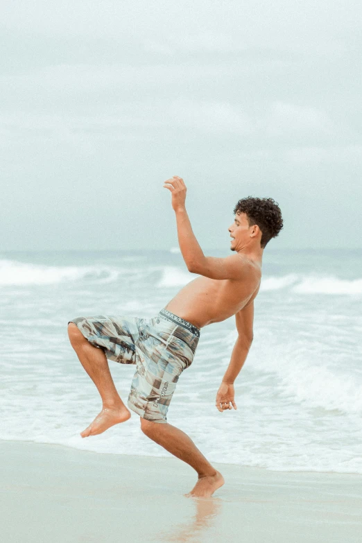 a man standing on top of a beach next to the ocean, arabesque, teen boy, doing a sassy pose, origin jumpworks, wears shorts