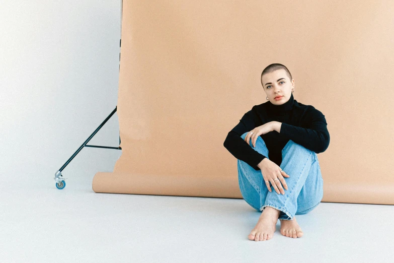 a woman sitting on the ground in front of a backdrop, trending on pexels, antipodeans, buzz cut, portrait sophie mudd, wearing turtleneck, full length portait