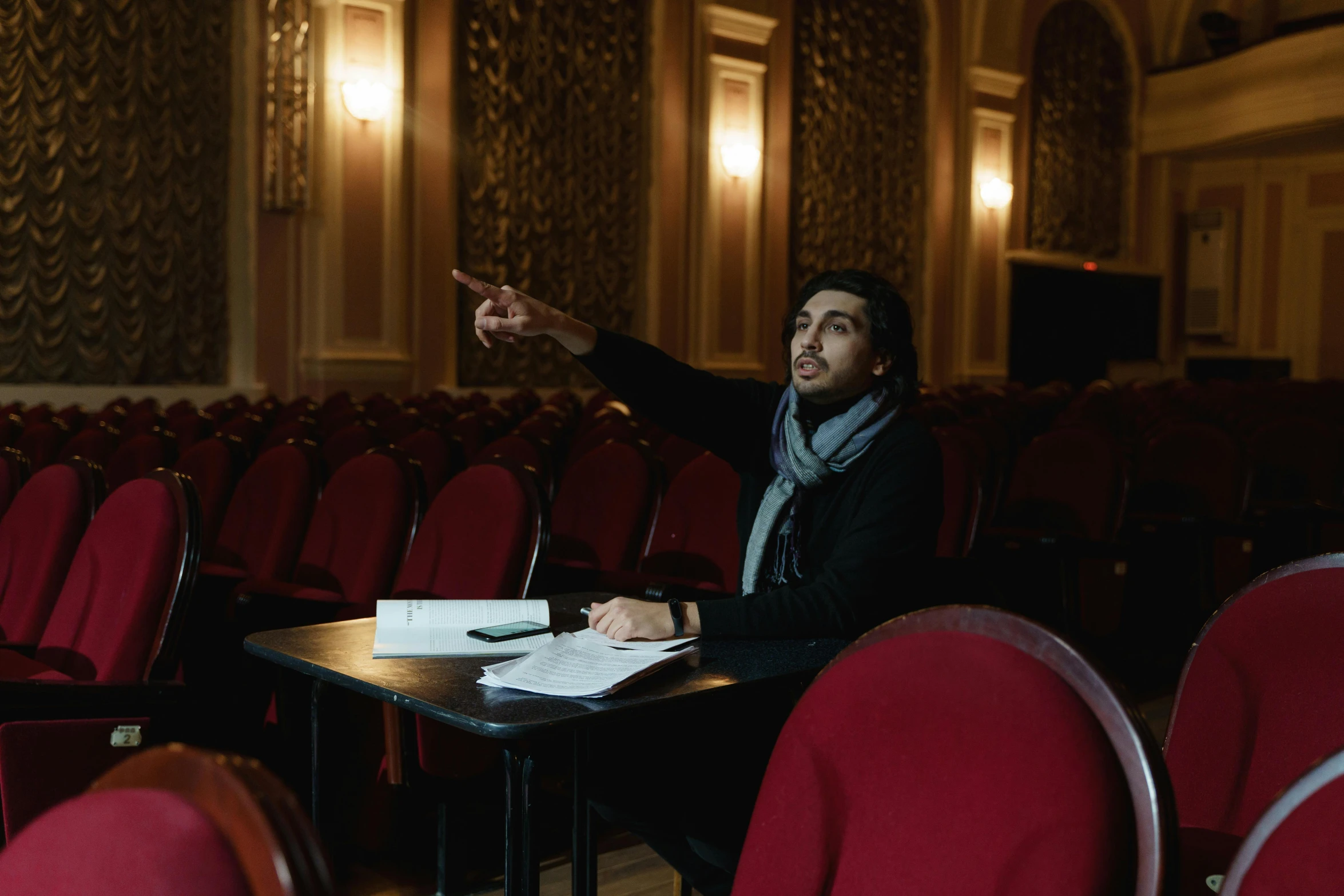 a man sitting at a table pointing at something, unsplash, academic art, theater, sam nassour, inside a grand, looking towards camera