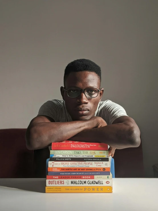 a man sitting at a table with a stack of books, by Lily Delissa Joseph, pexels contest winner, black arts movement, model posing, with glasses on, adut akech, an olive skinned