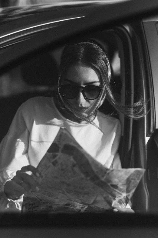 a woman sitting in a car reading a newspaper, a black and white photo, by Sergio Burzi, pexels contest winner, olivia culpo, wearing shades, cartography, performing