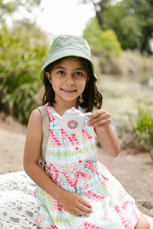 a little girl sitting on top of a blanket, white baseball hat, standing in a botanical garden, holding an ace card, modelling clay