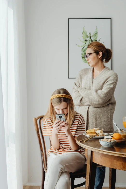 a woman and a little girl sitting at a table, trending on pexels, checking her phone, discontent, standing still, half image