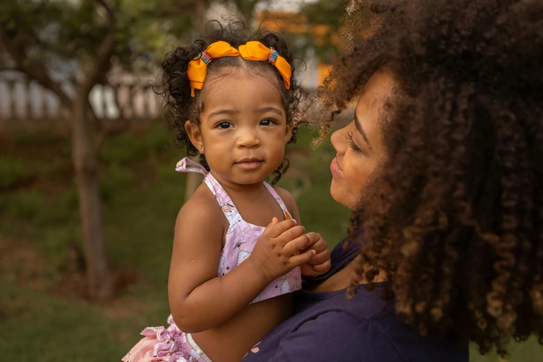 a woman holding a little girl in her arms, by Lily Delissa Joseph, pexels contest winner, curly afro, orange hair bow, ( brown skin ), 2 years old