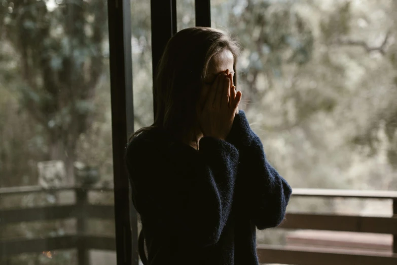 a woman standing in front of a window talking on a cell phone, inspired by Elsa Bleda, trending on pexels, woman crying, wearing a dark sweater, anorexic figure, closed hands