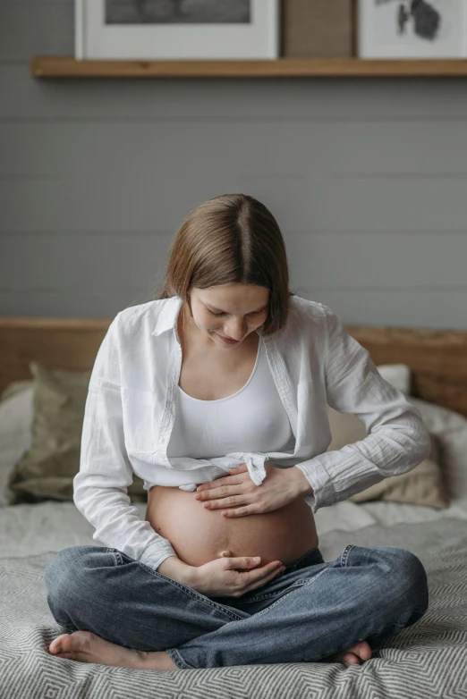 a pregnant woman sitting on a bed holding her stomach, pexels contest winner, covid-19 as a human, profile image, australian, white shirt and jeans