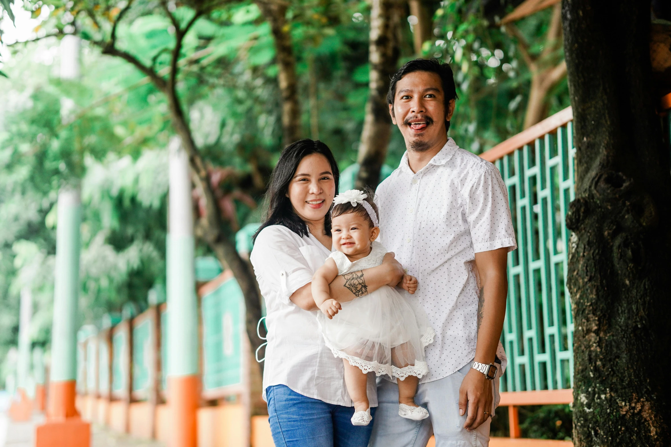 a man standing next to a woman holding a baby, a portrait, by Julia Pishtar, pexels contest winner, sumatraism, background image, of a family standing in a park, white, with mustache