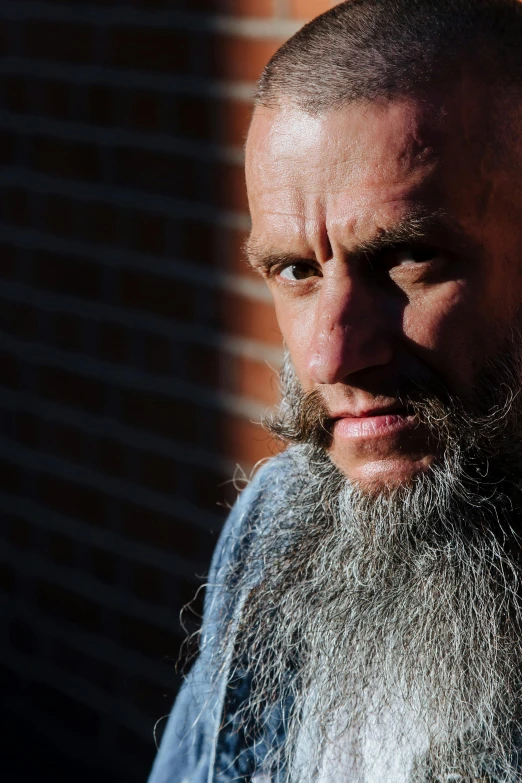 a man with a long beard standing in front of a brick wall, unsplash, hyperrealism, shaved head, tim booth, long grey hair, gangrel