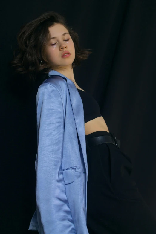 a woman standing in front of a black backdrop, an album cover, by irakli nadar, unsplash, realism, pale blue outfit, cropped shirt with jacket, kiko mizuhara, finn wolfhard