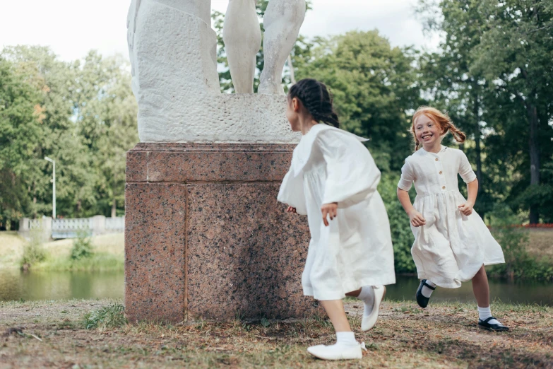 a couple of little girls standing next to a statue, a statue, inspired by Thomas Stothard, pexels contest winner, socialist realism, girl is running, wearing white cloths, stålenhag, at the park