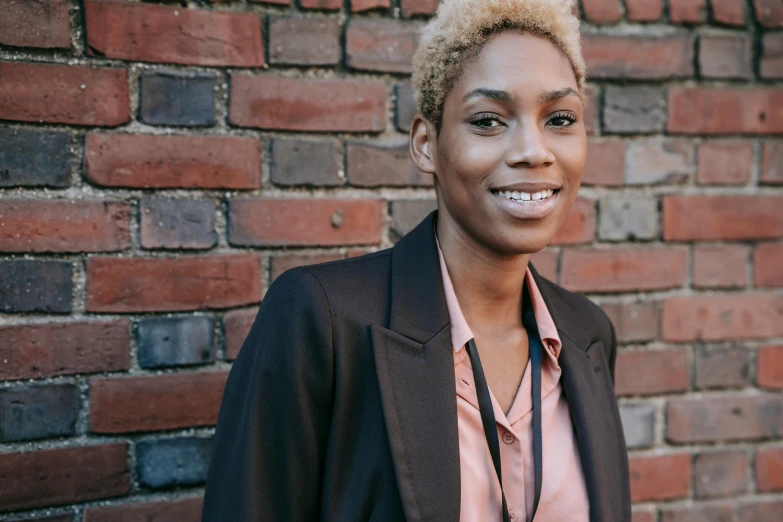 a woman standing in front of a brick wall, brown skinned, professional image, short blonde afro, thumbnail