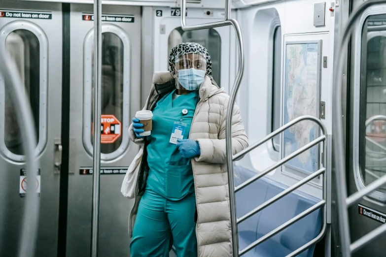 a woman standing on a subway holding a cup of coffee, by Meredith Dillman, pexels contest winner, happening, surgical gown and scrubs on, costume with blue accents, ny, 🚿🗝📝