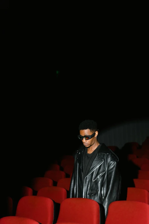 a man standing in front of a row of red chairs, an album cover, unsplash, realism, 2 1 savage, wearing shiny black goggles, sitting in a movie theater, a man wearing a black jacket