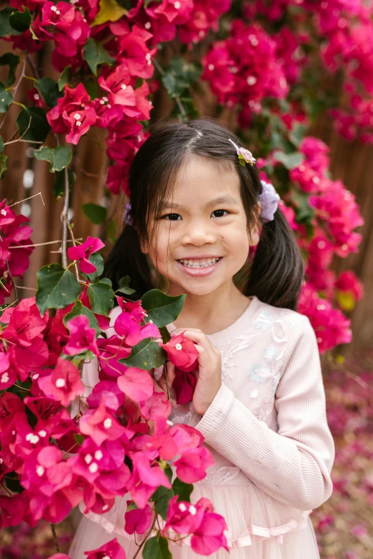 a little girl standing in front of a bush of pink flowers, while smiling for a photograph, katherine lam, bougainvillea, portrait image