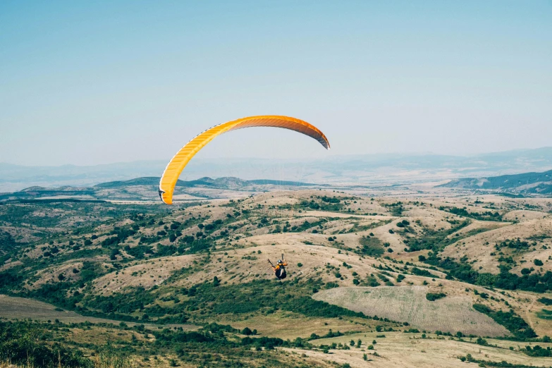 a person paragliding in the mountains on a sunny day, by Peter Churcher, unsplash contest winner, hurufiyya, hills in the background, ochre, victoria siemer, retro vibe