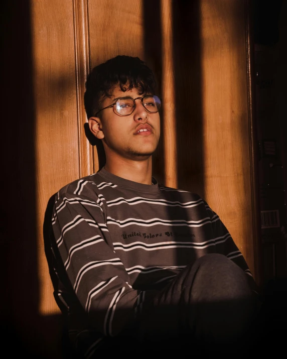 a young man wearing glasses sitting in a chair, an album cover, inspired by Ion Andreescu, trending on unsplash, sumatraism, lgbt, proud looking away, stripes, highly reflective