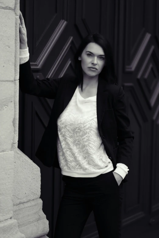 a black and white photo of a woman leaning against a wall, inspired by Luis Paret y Alcazar, business clothes, yennefer, wearing jacket, intricate fashion clothing