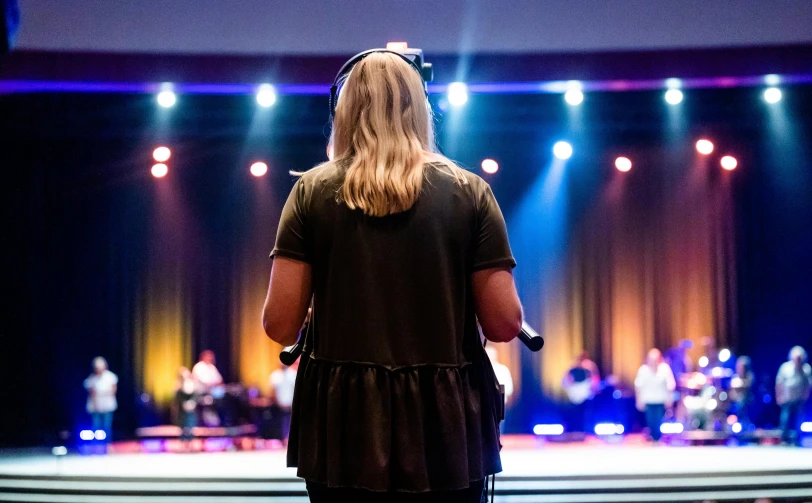 a woman standing in front of a stage with a microphone, unsplash, church background, from the back, student, realistic »