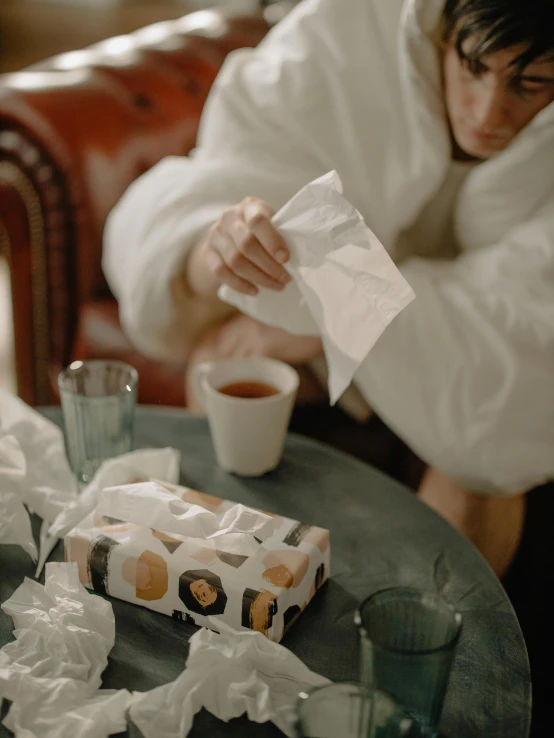 a man in a bathrobe sitting at a table with a box of tissues and a cup of coffee, by Emma Andijewska, pexels contest winner, ignant, made of lab tissue, people crying, holiday season