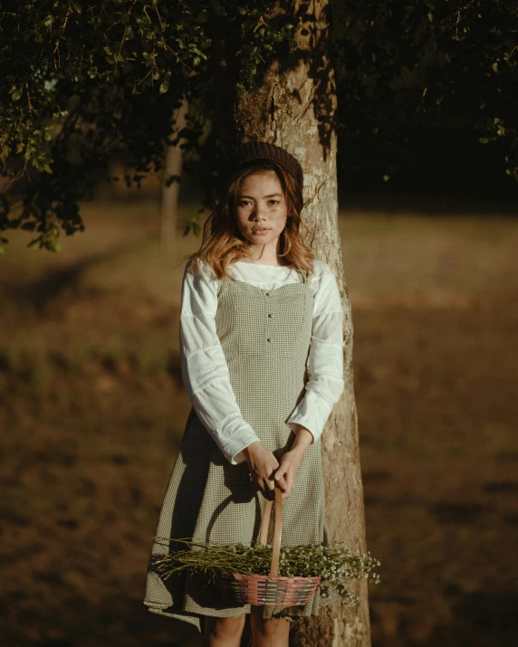 a woman standing next to a tree holding a basket, inspired by Camille Corot, pexels contest winner, renaissance, wearing overalls, ( ( theatrical ) ), hailee steinfeld, white apron