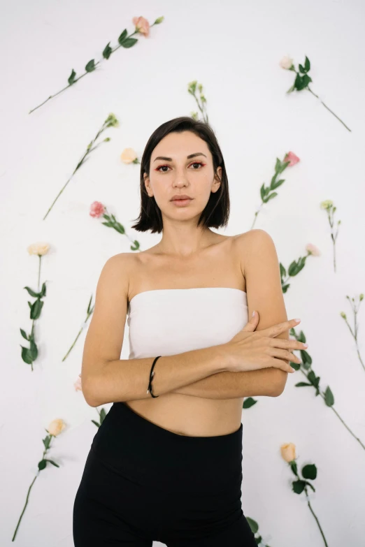 a woman standing in front of a wall with flowers, an album cover, inspired by Ilka Gedő, trending on pexels, wearing a cropped black tank top, in white room, alexandria ocasio - cortez, portrait pose