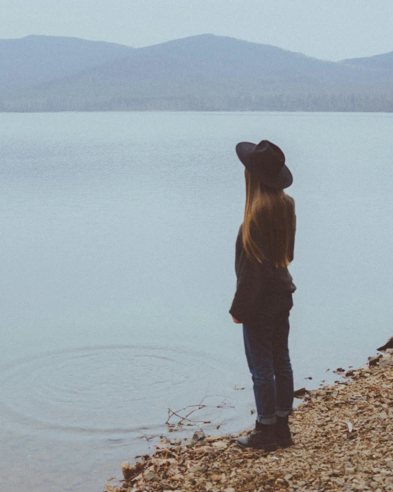 a woman standing on a beach next to a body of water, in the middle of a lake, shy looking down, androgynous person, looking off into the distance