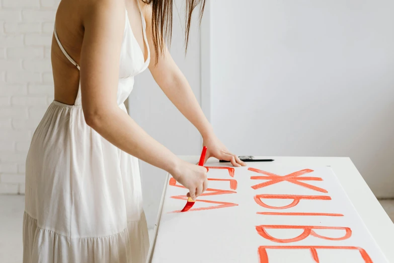 a woman in a white dress is cutting a sign, by Julia Pishtar, trending on pexels, letterism, on a white table, light red and orange mood, detailed acrylic, making out